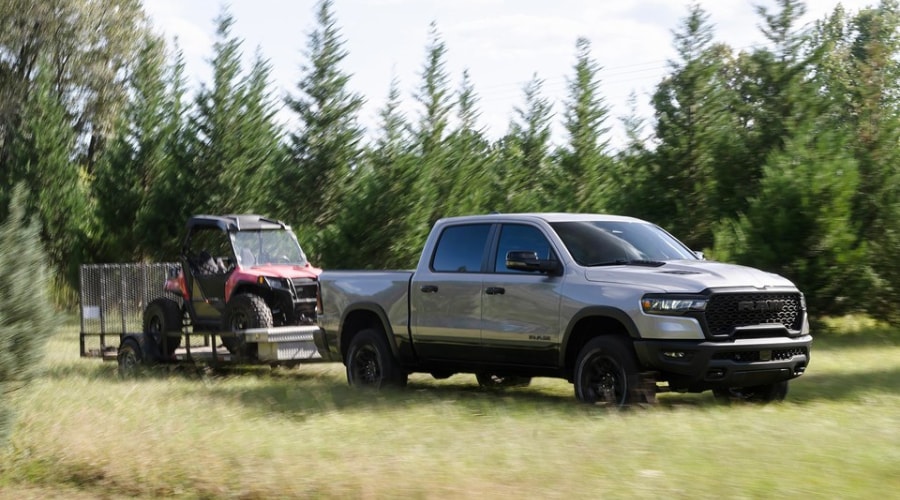 Silver 2026 Ram 1500 Crew Cab towing a utility trailer loaded with a red UTV through a field, demonstrating Trailer Auto Hitch Assist and towing power at Route 18 CJDR near East Brunswick and Monroe, NJ.