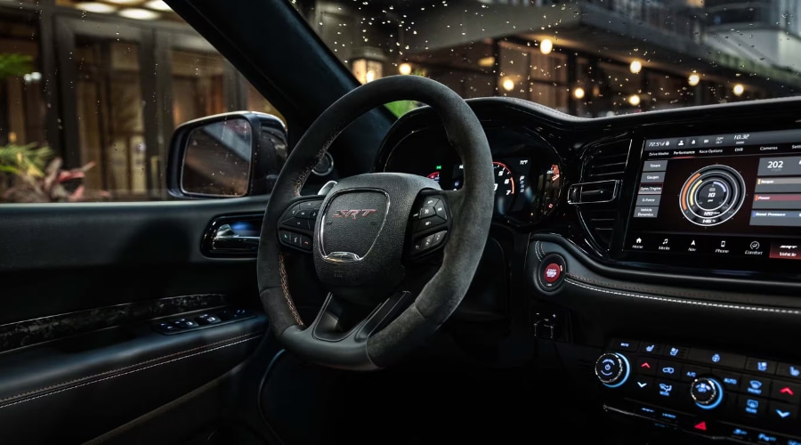 Driver cockpit view inside a 2026 Dodge Durango showing a suede-wrapped steering wheel, digital drive mode touchscreen, and rain-streaked side window during a stormy evening drive in East Brunswick NJ.