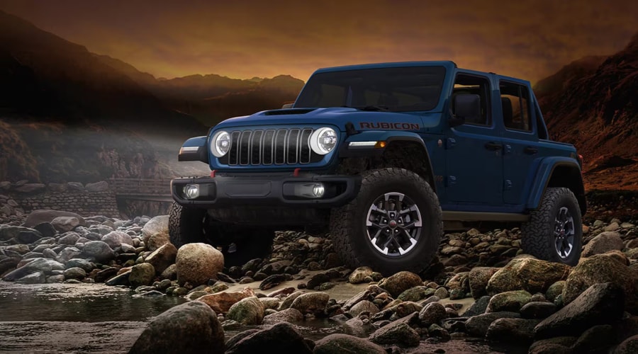 Front three-quarter view of a dark blue 2026 Jeep Wrangler Rubicon with illuminated LED halo headlights parked on a rocky autumn landscape, demonstrating off-road 4x4 capability and pre-winter maintenance at Route 18 Autos in East Brunswick, NJ.