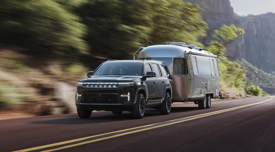 2026 Jeep Grand Wagoneer towing a silver travel trailer along a highway, demonstrating 9,800 lbs max towing capacity near East Brunswick, NJ.