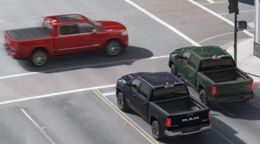 Three different 2026 Ram 1500 pickup trucks in red, dark blue, and dark green driving through a local intersection, representing the diverse inventory options ready for a test drive at Route 18 Autos in East Brunswick, New Jersey.
