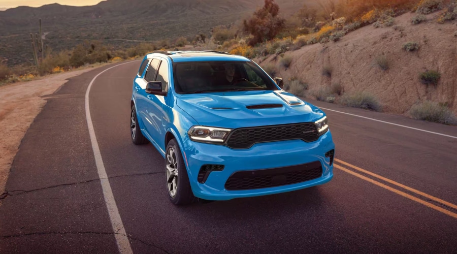 Front three-quarter view of a bright blue 2026 Dodge Durango driving along a winding two-lane road with headlights on, showcasing all-wheel drive traction and handling stability for East Brunswick NJ commuters.