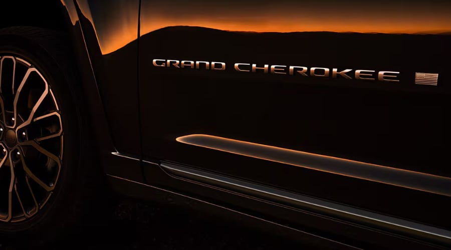 Close-up side profile of a dark metallic 2026 Jeep Grand Cherokee showing the chrome Grand Cherokee door badge, American flag emblem, and alloy wheel design at Route 18 Autos in East Brunswick, NJ.