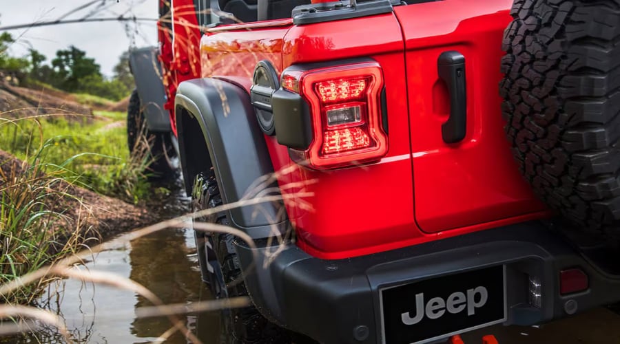 Rear view of a bright red 2026 Jeep Wrangler navigating a damp trail with an illuminated LED taillight and rugged all-terrain spare tire, showcasing autumn capability and pre-winter maintenance at Route 18 Autos in East Brunswick, NJ.