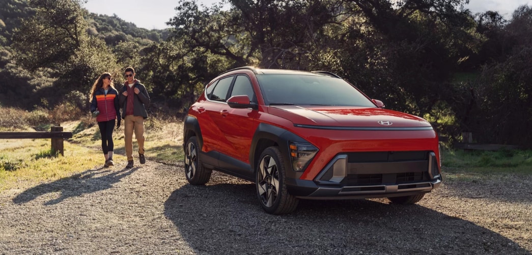 Orange 2026 Hyundai Kona parked in forested area with couple walking next to it.