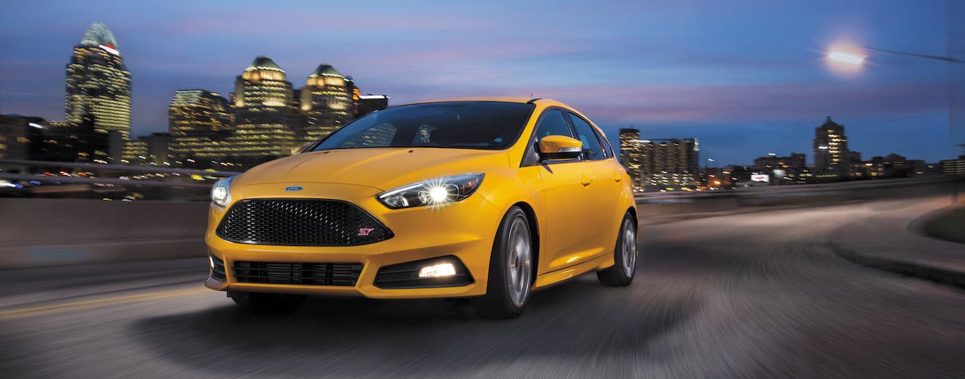 A yellow 2016 Ford Focus ST is shown driving past a city at night.
