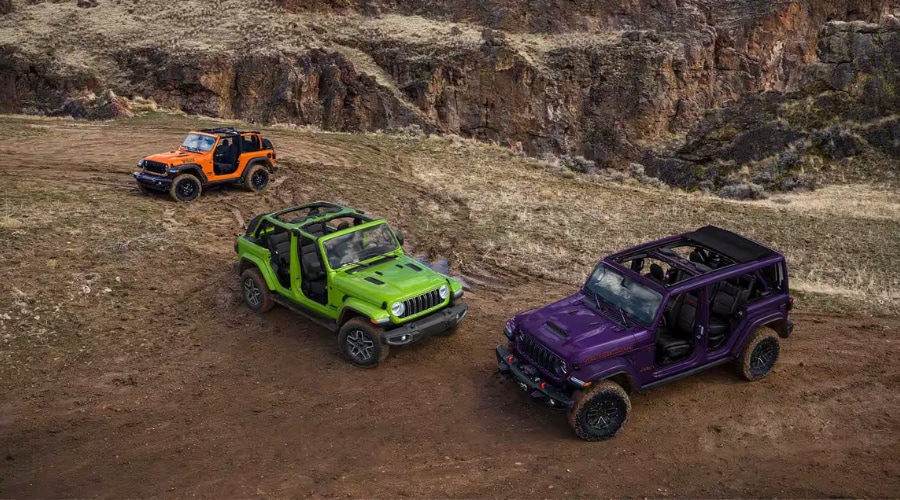 Three 2026 Jeep Wrangler models including a purple Rubicon, bright green Unlimited, and orange 2-door Willys parked on a rugged dirt trail near Morristown NJ demonstrating open-air configurations with doors and roofs removed