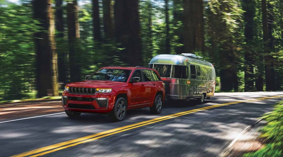 Red 2027 Jeep Grand Cherokee SUV towing a silver camper trailer on a scenic highway, demonstrating its best-in-class 6,200-pound maximum towing capacity available at Salerno Duane Chrysler Jeep Dodge Ram in New Jersey.