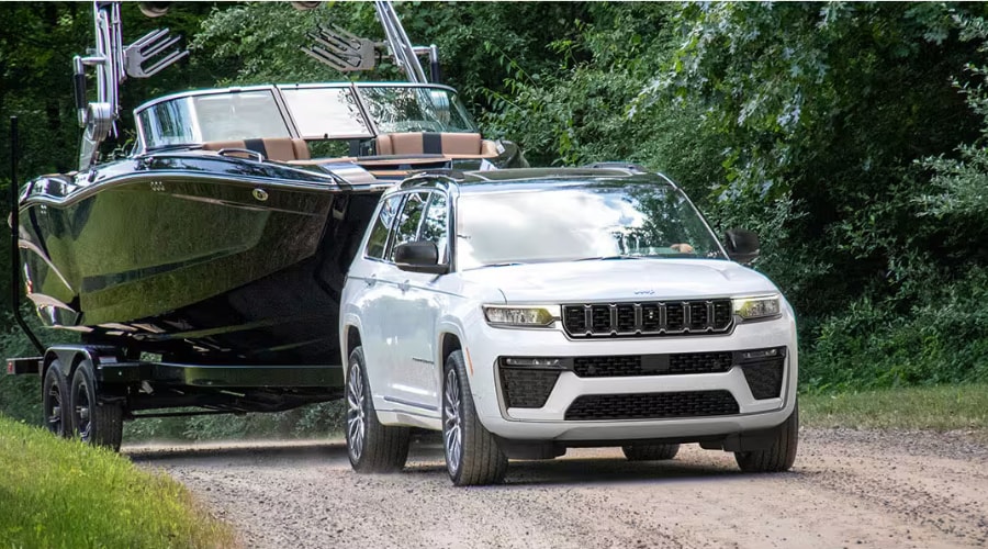 A 2026 Jeep Grand Cherokee demonstrating its high-torque towing capacity by pulling a large boat on a trailer down a gravel road near Old Bridge New Jersey