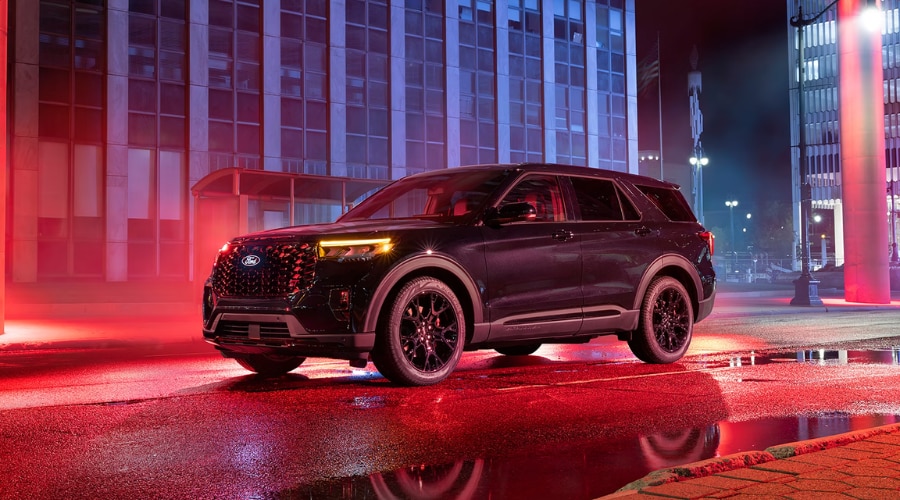 Side three-quarter view of a dark metallic 2026 Ford Explorer parked on a wet city street at night under red neon reflections, highlighting illuminated amber LED running lights, black alloy wheels, and all-weather traction capability for rainy commutes near Morristown and Summit, NJ.