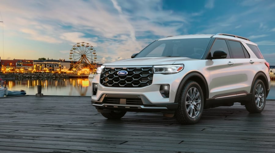 Three-quarter front angle of a 2026 Ford Explorer in Shiny Gray parked on a wooden boardwalk pier at sunset, showcasing its bold honeycomb front grille and LED headlights against a harbor background with an illuminated Ferris wheel.