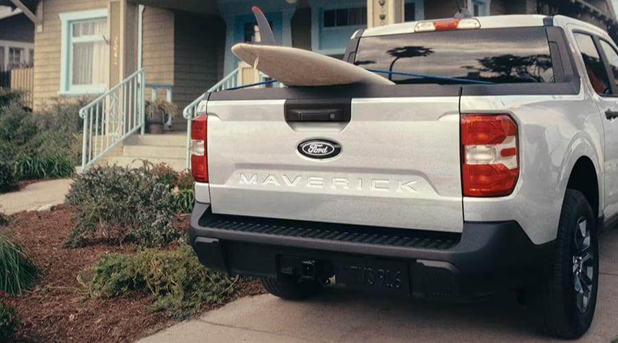 Rear view of a silver Ford Maverick compact pickup truck parked in a suburban driveway in Union County NJ, featuring a surfboard loaded in the bed for weekend adventures.