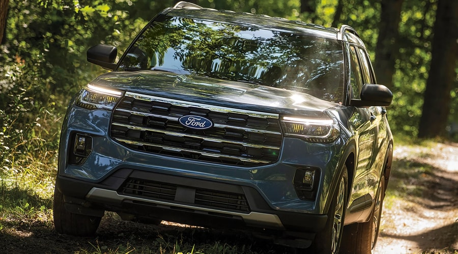 Front view of a slate blue 2027 Ford Explorer driving along a sunlit, wooded dirt trail, showcasing its updated chrome-accented front grille, signature LED headlights, and silver skid plate at Salerno Duane Ford near Summit, NJ.