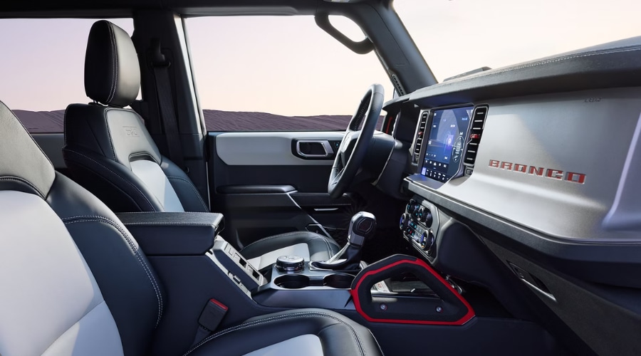 Spacious interior front cabin of a 2026 Ford Bronco showing two-tone black and light grey leather seats, central SYNC 4 touchscreen display, rotary G.O.A.T. Modes dial, and red-accented passenger grab handle, available at Salerno Duane Ford near Summit and Morristown, NJ.