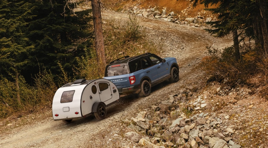 A slate blue 2026 Ford Bronco Sport 4x4 towing a silver teardrop camper trailer uphill on a steep gravel mountain trail surrounded by pine trees, demonstrating its 2,200 lb towing capability for weekend camping trips from Summit and Morristown, NJ.