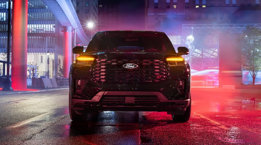 Front view of a dark 2027 Ford Explorer ST parked on a wet city street at night, showcasing its updated honeycomb front grille, red ST emblem, and glowing amber LED headlight signature at Salerno Duane Ford near Summit, NJ.