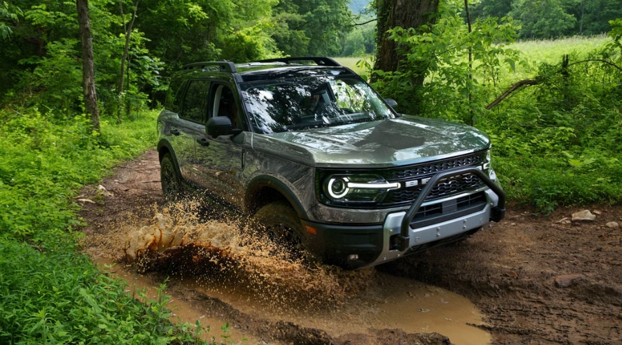 A dark gray 2026 Ford Bronco Sport driving through a muddy puddle on a lush green woodland trail, highlighting standard 4x4 capability and reliable wet-road handling for drivers in Summit, NJ, and Morris County.