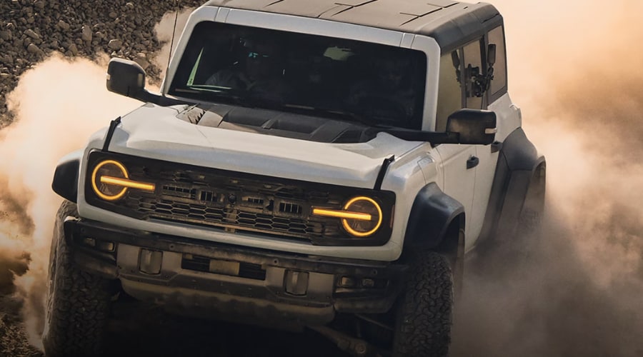 Front three-quarter view of a white 2026 Ford Bronco Raptor with illuminated amber halo headlights charging through a thick dust cloud, demonstrating extreme off-road power for drivers near Summit, NJ.
