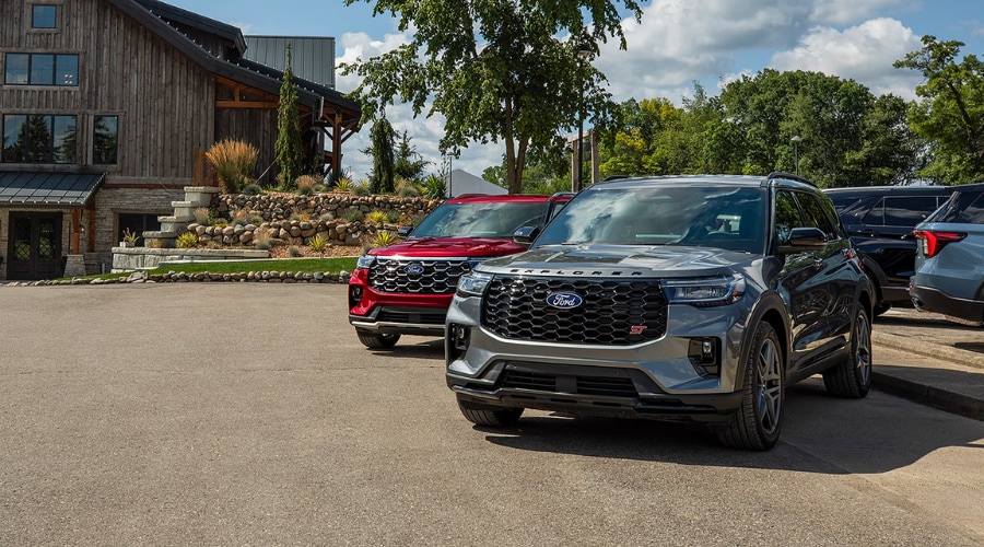 Front three-quarter view of a metallic grey 2026 Ford Explorer ST parked alongside a red Ford Explorer in a paved lot outside a suburban lodge, highlighting 3-row family SUV trim options and all-weather capability available near Summit and Morristown, NJ.