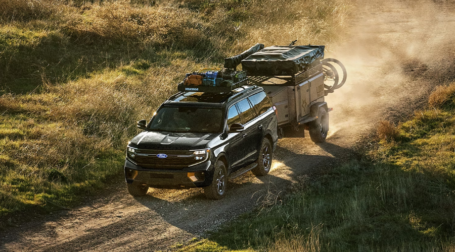 Black 2027 Ford Expedition Tremor towing an overland camper trailer and carrying a loaded roof basket along a dirt road, demonstrating up to 9,600 lbs towing capacity at Salerno Duane Ford near Paramus, NJ.