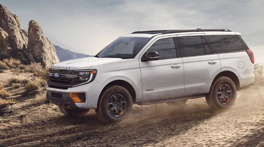 White 2027 Ford Expedition Tremor full-size off-road SUV with orange front recovery hooks, black grille, dark alloy wheels, and all-terrain tires driving on a dusty trail at Salerno Duane Ford near Paramus, NJ.