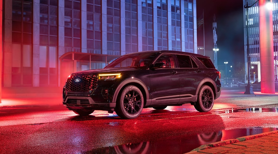 Side-profile view of a dark metallic 2027 Ford Explorer Platinum parked on a wet city street at night under vivid red neon lighting, featuring gloss-black alloy wheels, red brake calipers, and an illuminated Ford front badge at Salerno Duane Ford near Summit, NJ.