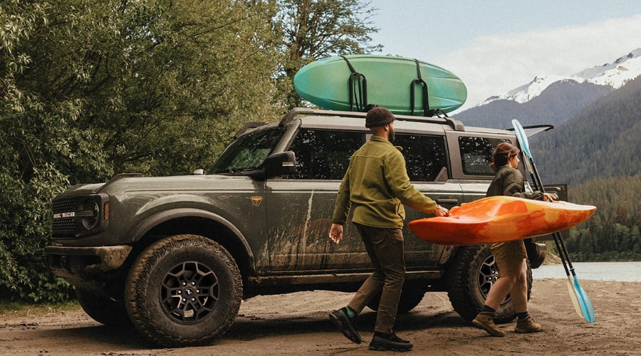 Mud-splattered 2026 Ford Bronco Badlands parked near a lake shore as two outdoor enthusiasts unload kayaks, showing off-road capability for Union County and Morris County weekend trail trips.