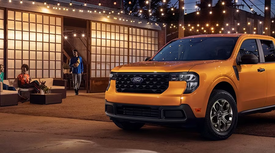 Front view of a yellow-orange 2026 Ford Maverick compact truck parked outside an illuminated venue at dusk in North Jersey, ideal for daily commutes across Union and Morris County