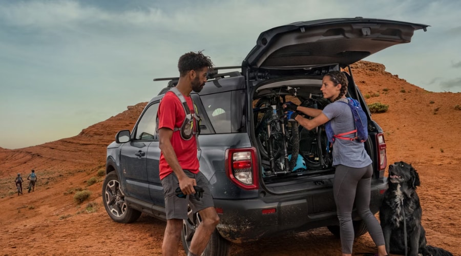 A couple loading two upright mountain bikes into the rear cargo space of a slate blue 2026 Ford Bronco Sport with an open liftgate and safari-style roof, demonstrating versatile interior cargo capacity for outdoor adventures near Watchung Reservation and Union County, NJ.