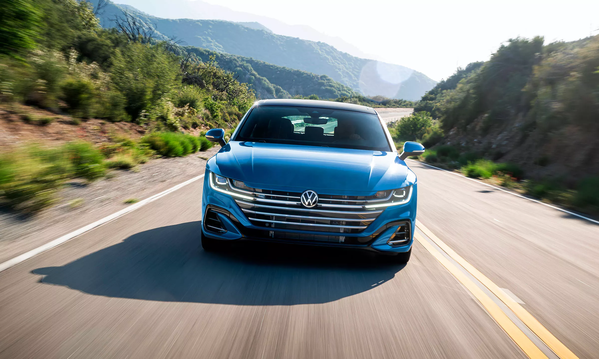 A front view of Arteon in Kingfisher Blue Metallic coming out of a curve on a two-lane road with mountains behind it.