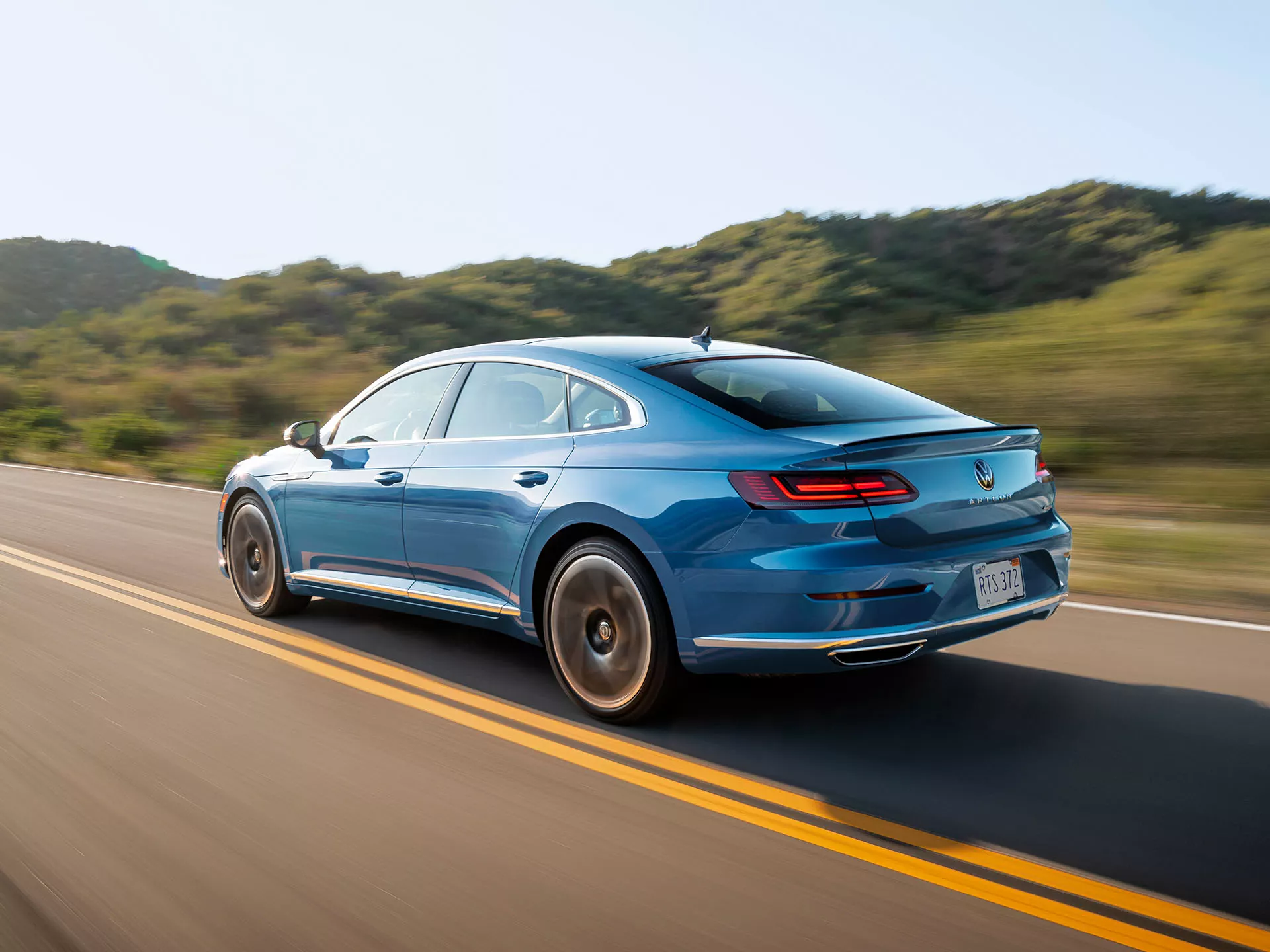 An Arteon shown in Kingfisher Blue Metallic accelerates along a two-lane road lined with hills. 