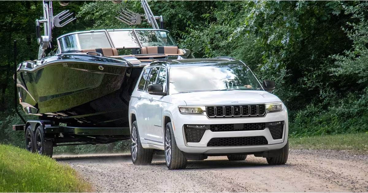  A white SUV, a 2026 Jeep Grand Cherokee, towing a sleek black wakeboat on a tandem-axle trailer along a dirt road surrounded by dense green foliage. The SUV features a distinctive front grille, roof rails, and multi-spoke rims, while the boat has a pointed bow, tan seating, and a wakeboard tower. The scene conveys outdoor adventure and recreational boating.