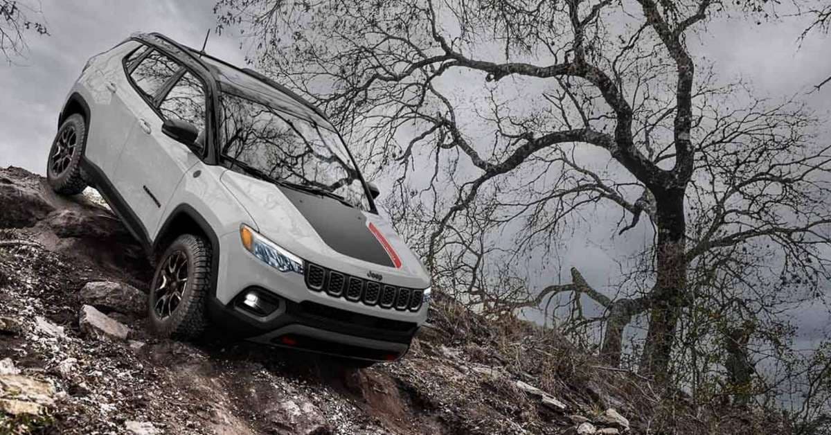 A white Jeep Compass Trailhawk navigating a steep, rocky, and muddy incline under an overcast sky. The SUV features a black roof, black hood accents with a red stripe, red tow hooks, and rugged all-terrain tires. Its headlights and fog lights are illuminated, emphasizing its off-road capabilities. A large, bare tree with gnarled branches stands in the background, adding to the rugged and adventurous atmosphere.