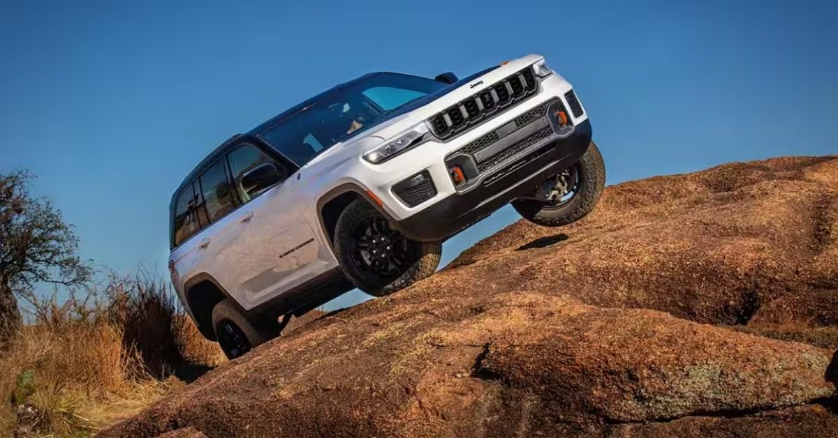 A white Jeep Grand Cherokee Trailhawk navigating rugged off-road terrain with large reddish-brown rocks. The vehicle is angled upward, with its front left wheel elevated on a rock, showcasing its off-road capabilities. It features a seven-slot grille, red tow hooks, black accents, and off-road tires. The background includes sparse dry vegetation, a tree, and a clear blue sky, emphasizing an adventurous and rugged setting.