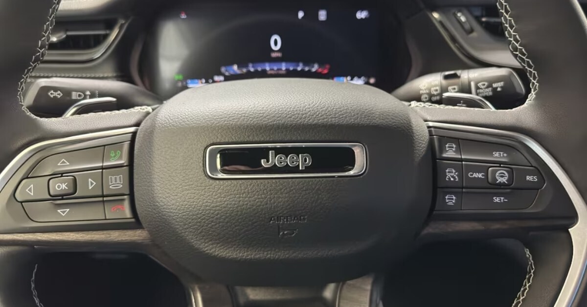 A close-up view of a 2026 Jeep Grand Cherokee's steering wheel and digital instrument cluster. The black leather steering wheel features white stitching, a metallic 