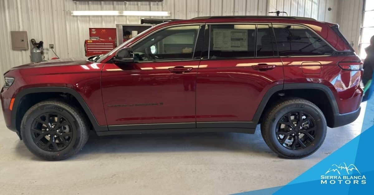 A side view of a dark red 2026 Jeep Grand Cherokee parked indoors in a garage or showroom. The SUV features black multi-spoke wheels, roof rails, and a glossy finish. The background includes a corrugated metal wall, a red toolbox, and a gas cylinder, indicating an automotive service or sales environment. A 