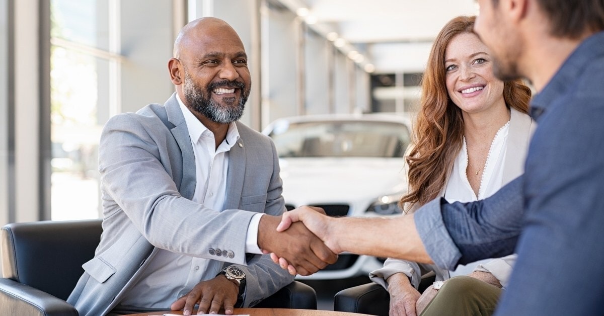 An indoor scene at a car dealership featuring a handshake between two men. The man on the left, wearing a light grey suit and smiling, shakes hands with a man in a dark blue shirt whose back is to the viewer. A woman with long reddish-brown hair, wearing a light-colored blazer, sits beside the man in the suit, smiling warmly. A dark table with documents is in the foreground, and a white car is visible in the well-lit background through large windows. The image conveys a professional and positive business interaction.