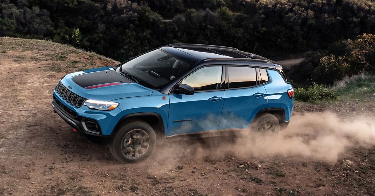 A blue Jeep Compass Trailhawk driving on a rugged dirt path, kicking up a large cloud of dust behind it. The SUV features a black roof, black hood decal with red accents, red tow hooks, and chunky off-road tires. The iconic seven-slot Jeep grille and 