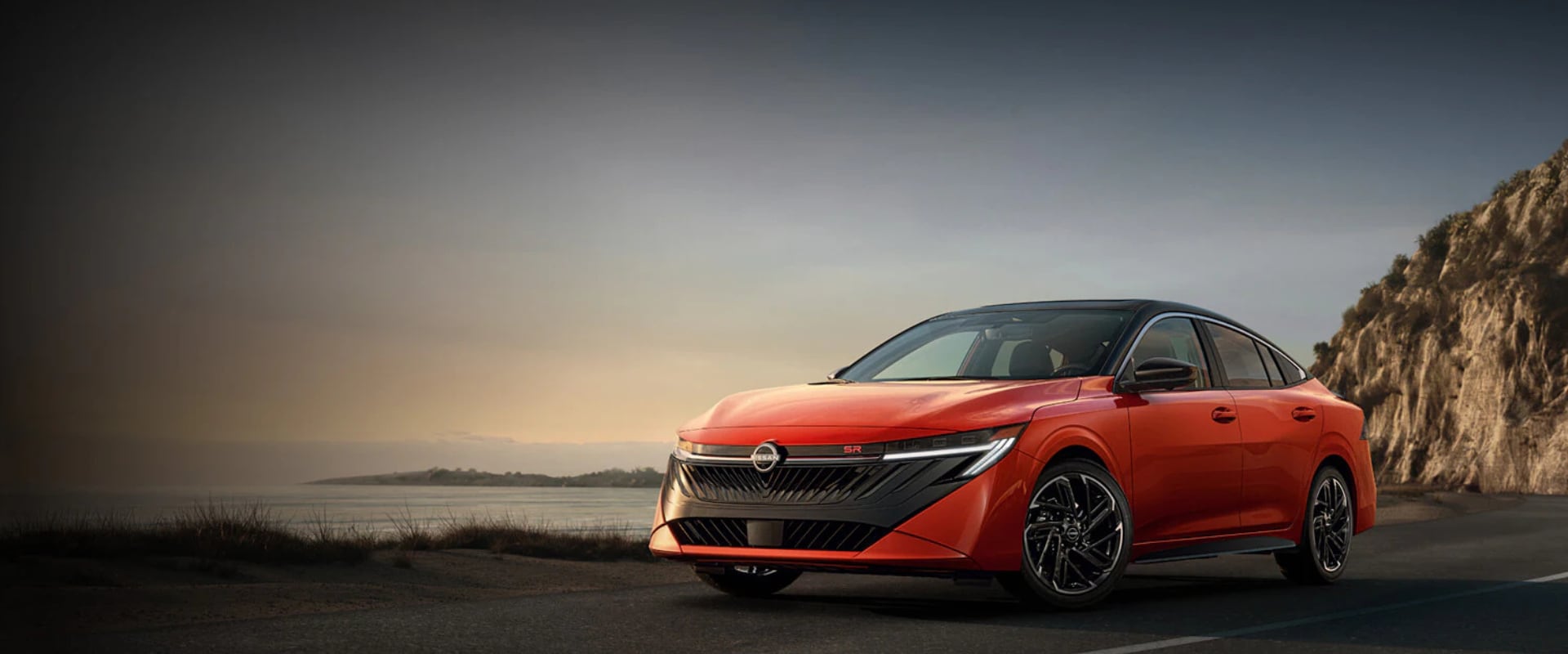 Exterior front angle image of a red-orange 2026 Nissan Sentra parked on a waterside cliff