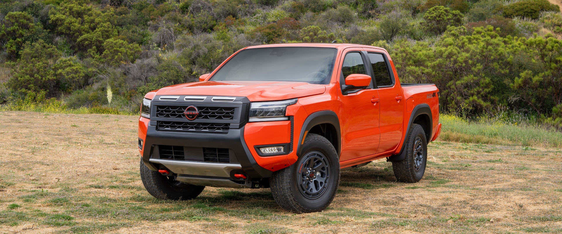 Front angled exterior image of an orange 2025 Nissan Frontier parked in an open field with trees behind it