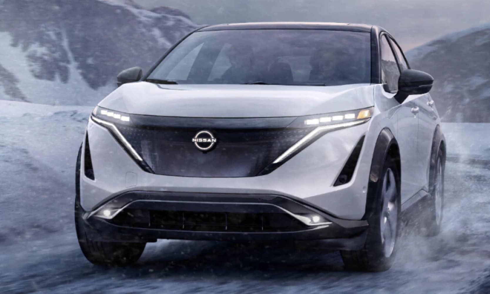 Closeup front-view image of a white 2025 Nissan ARIYA PLATINUM+ driving through a snowy mountain landscape