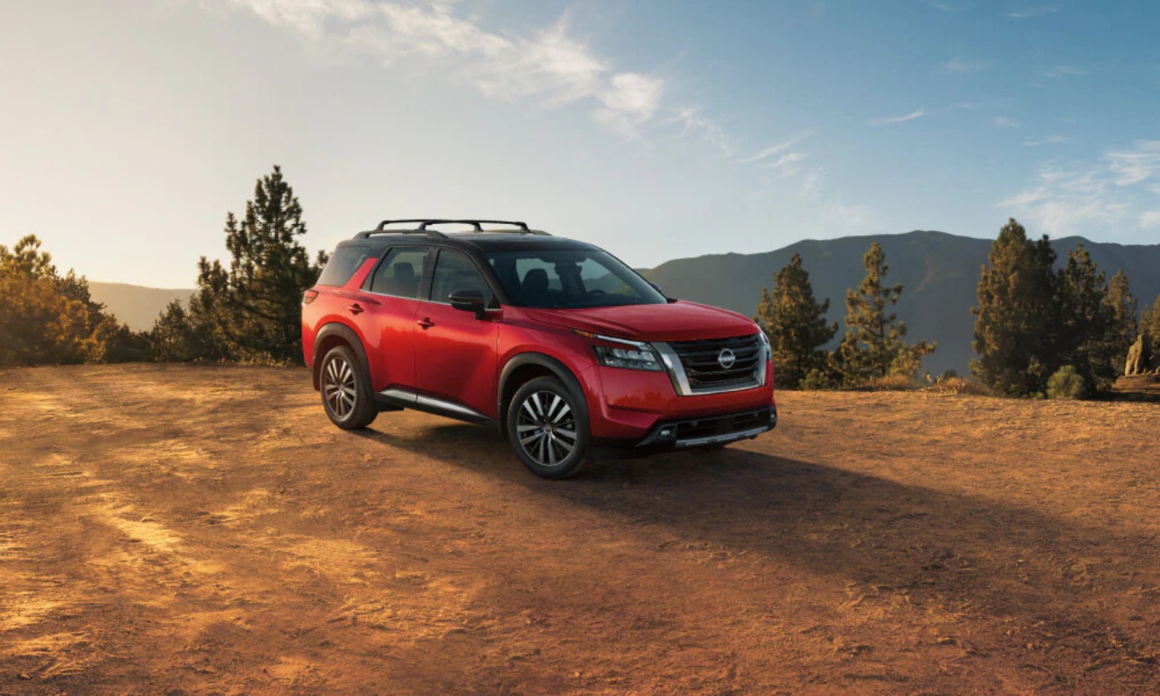 Exterior image of a red 2025 Nissan Pathfinder Platinum parked on a forest hill with mountains in the background