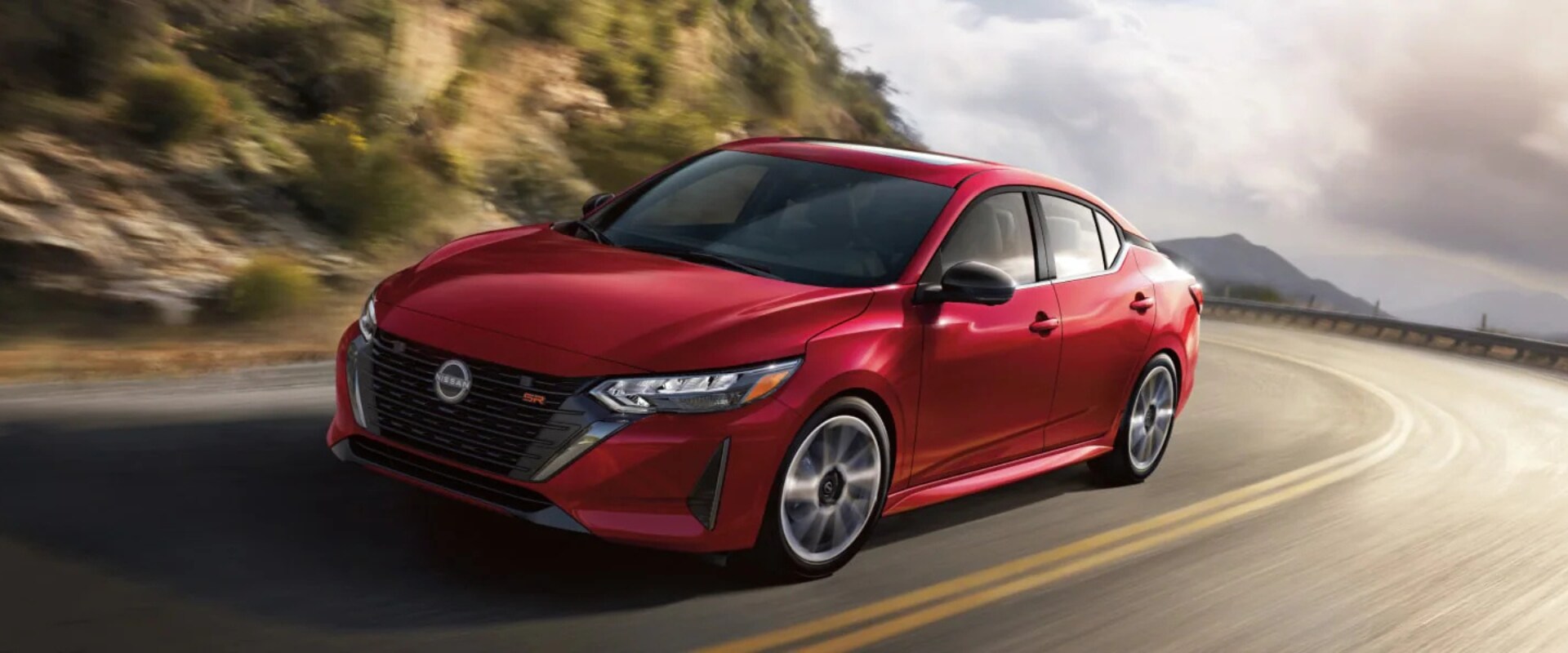 Exterior hero image of a red 2025 Nissan Sentra driving up a mountain road on a cloudy day