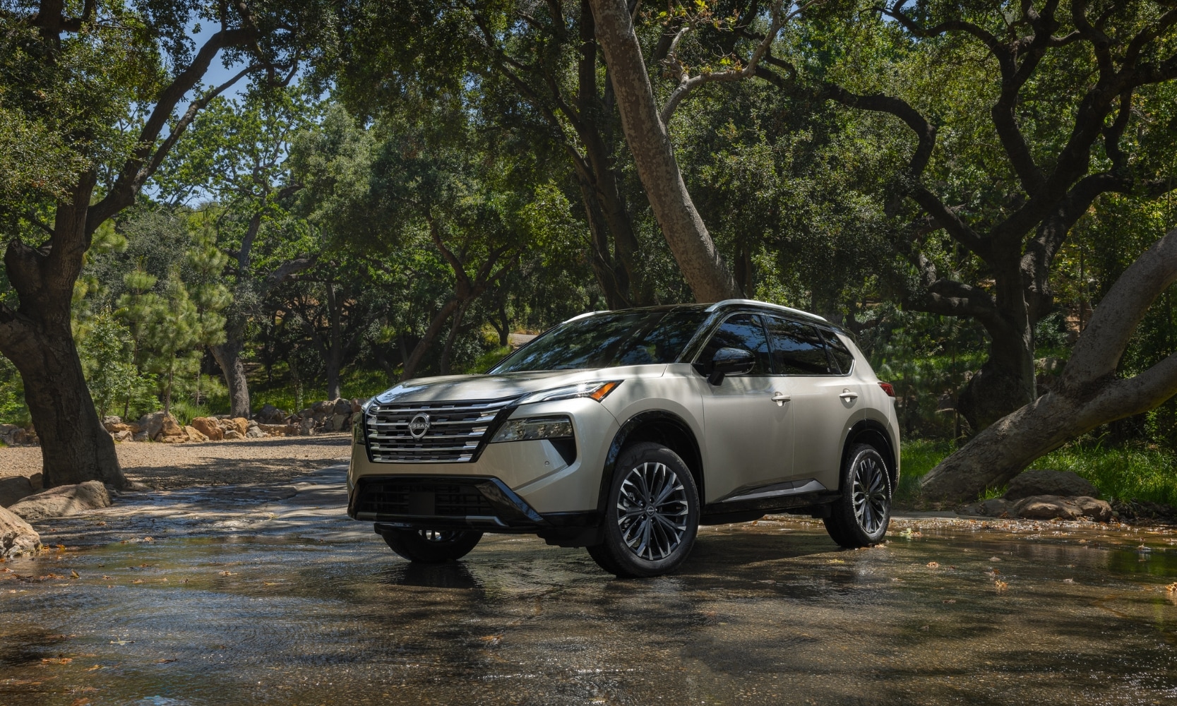 Exterior side angle image of a light beige 2024 Nissan Rogue driving through a rural park and shallow water