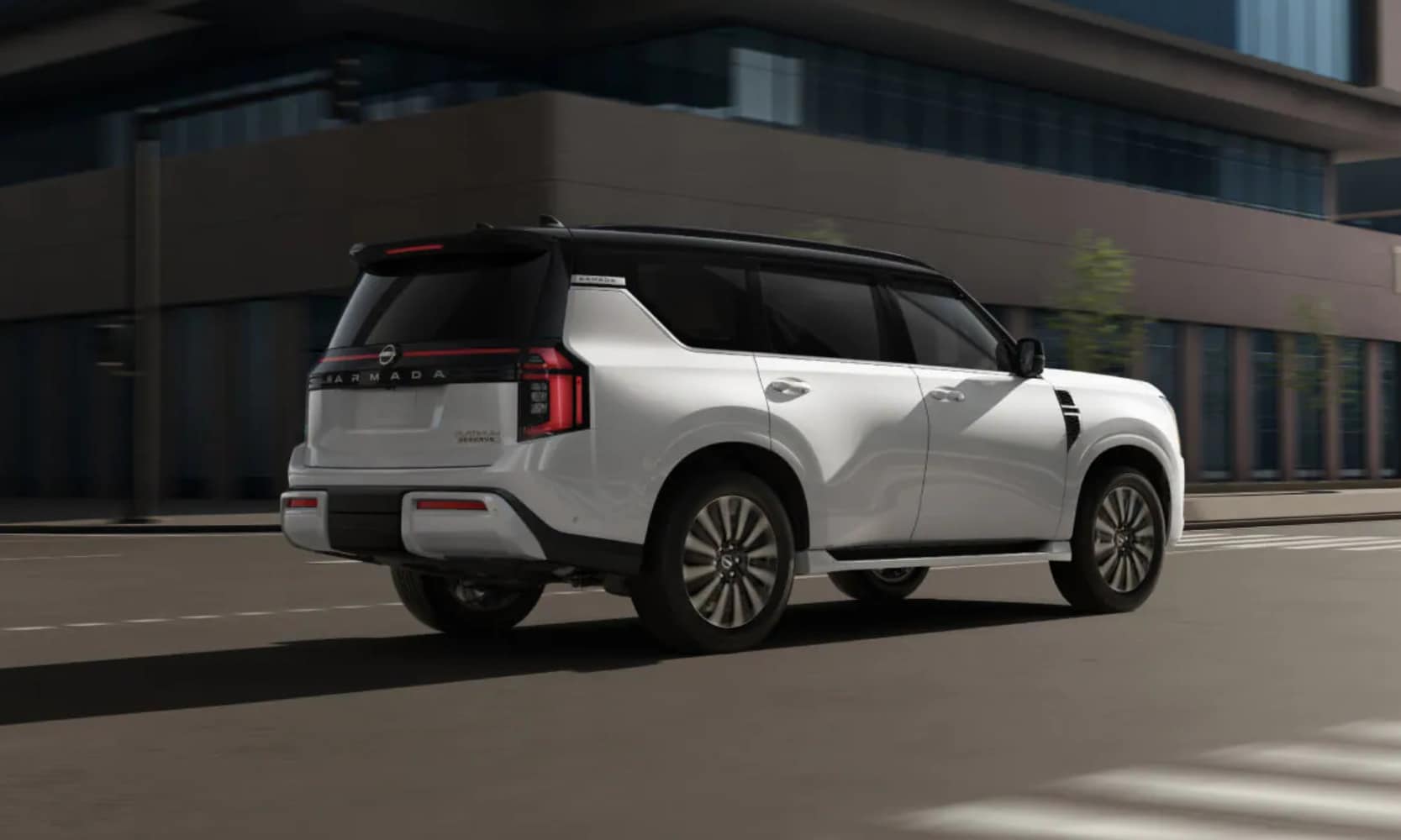 Exterior rear side image of a white 2025 Nissan Armada Platinum Reserve driving down a city street passing a building