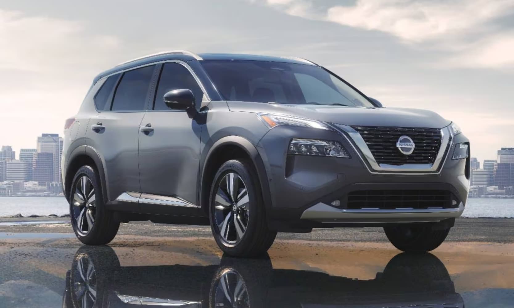 Exterior side angle image of a gray 2021 Nissan Rogue Platinum parked with skyscrapers and a lake in the background