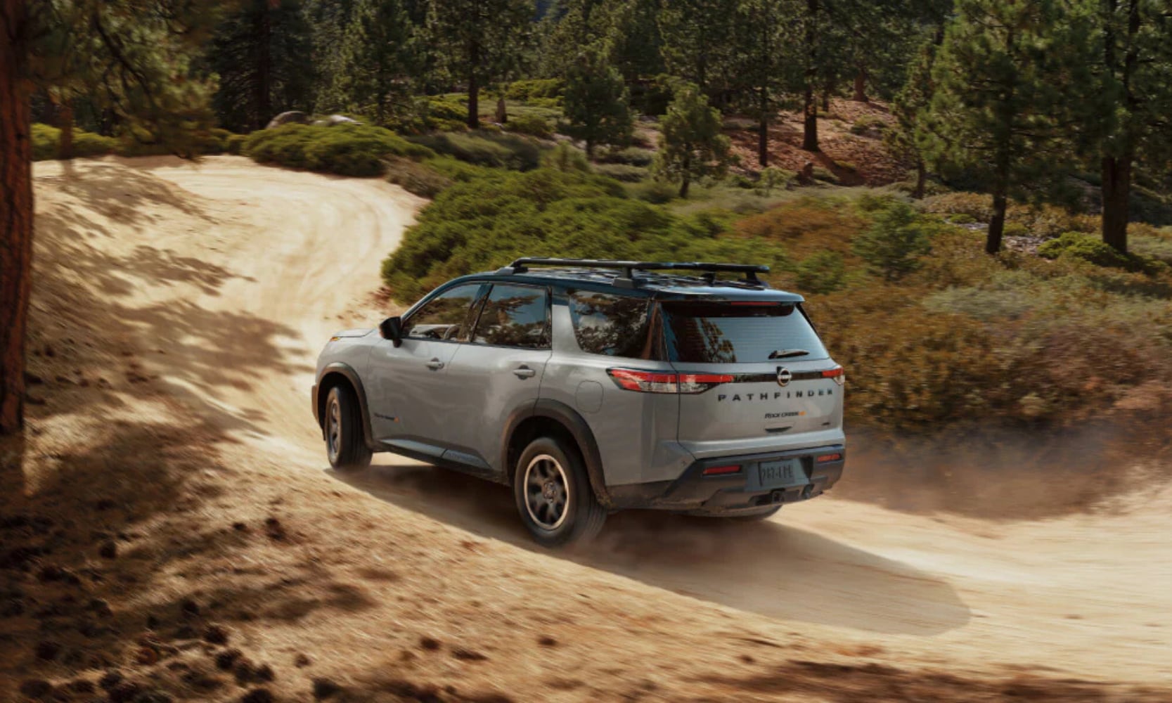 Exterior rear-view image of a gray 2025 Nissan Pathfinder Rock Creek driving up a dirt hill in a forest