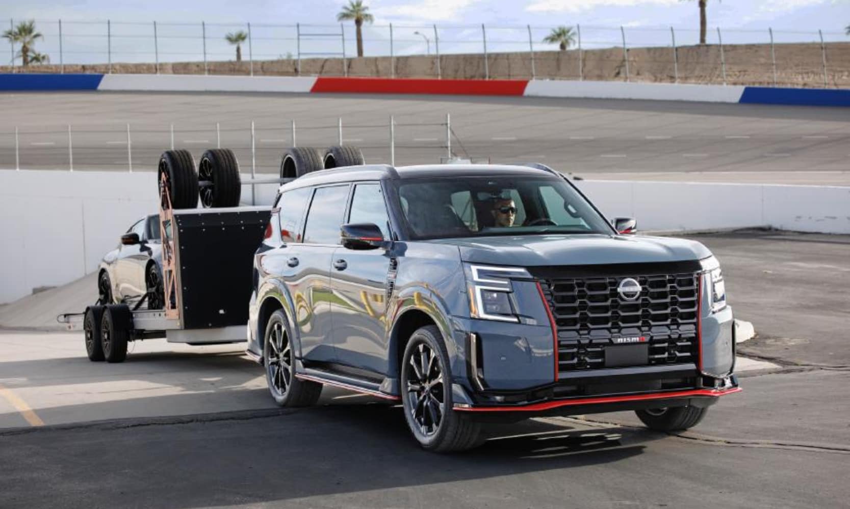 Exterior front angle image of a 2026 Nissan Armada NISMO towing a car on a trailer on a raceway track