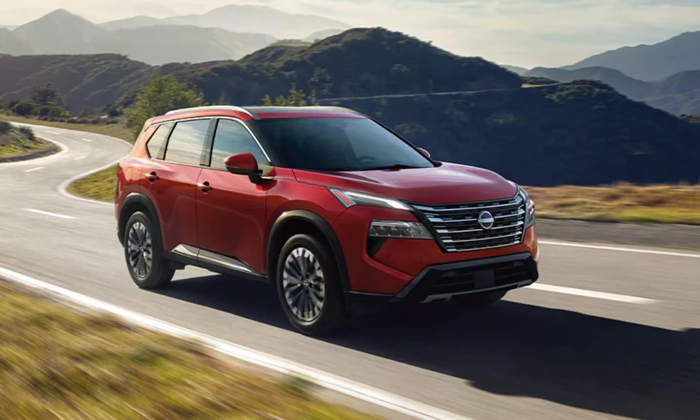 Exterior side image of a red 2025 Nissan Rogue Platinum driving on an open road with mountains in the background