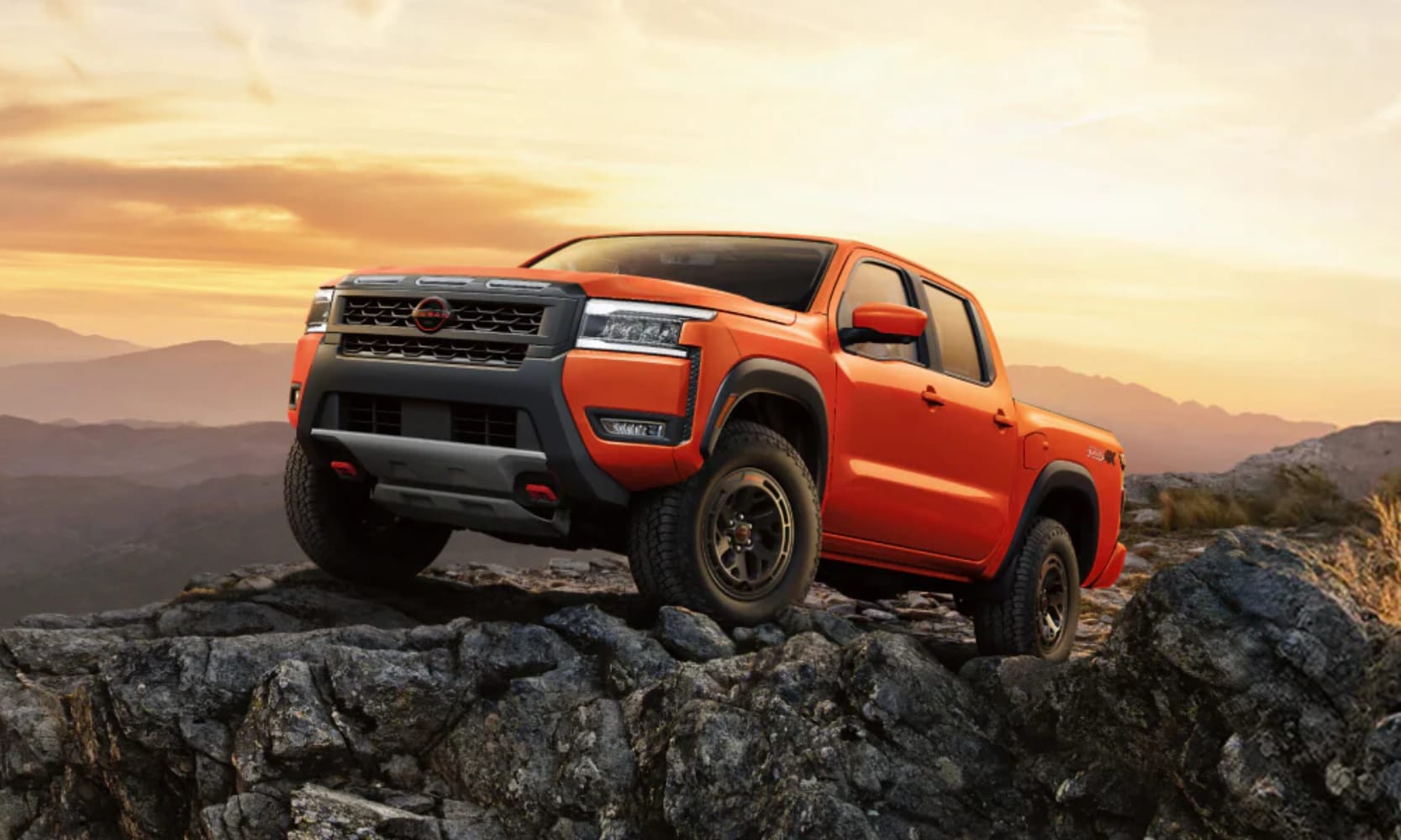 Exterior side angled image of an orange 2025 Nissan Frontier PRO-4X parked on a rocky mountain during sunset